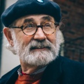An old man with gray beard, glasses and a hat looking to the horizon